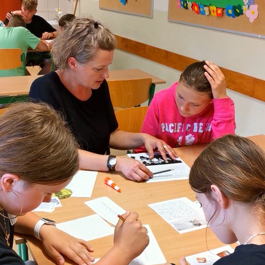 W klasie przy ławce szkolnej siedzi kobieta oraz troje uczniów, którzy wspólnie pochylają się nad rozłożonymi na stole kartkami papieru zawierającymi zadania do wykonania
