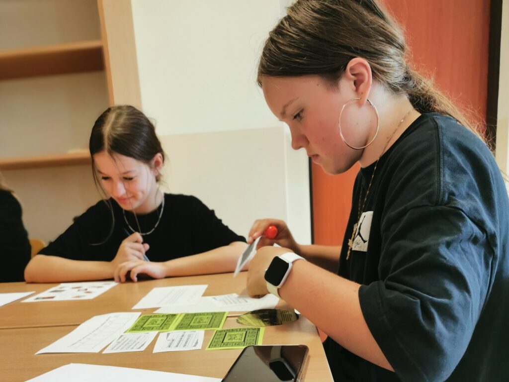 W klasie w ławce szkolnej siedzą dwie uczennice przyglądające się zadaniom umieszczonym na białych i żółtych kartkach ułożonych na blacie ławki