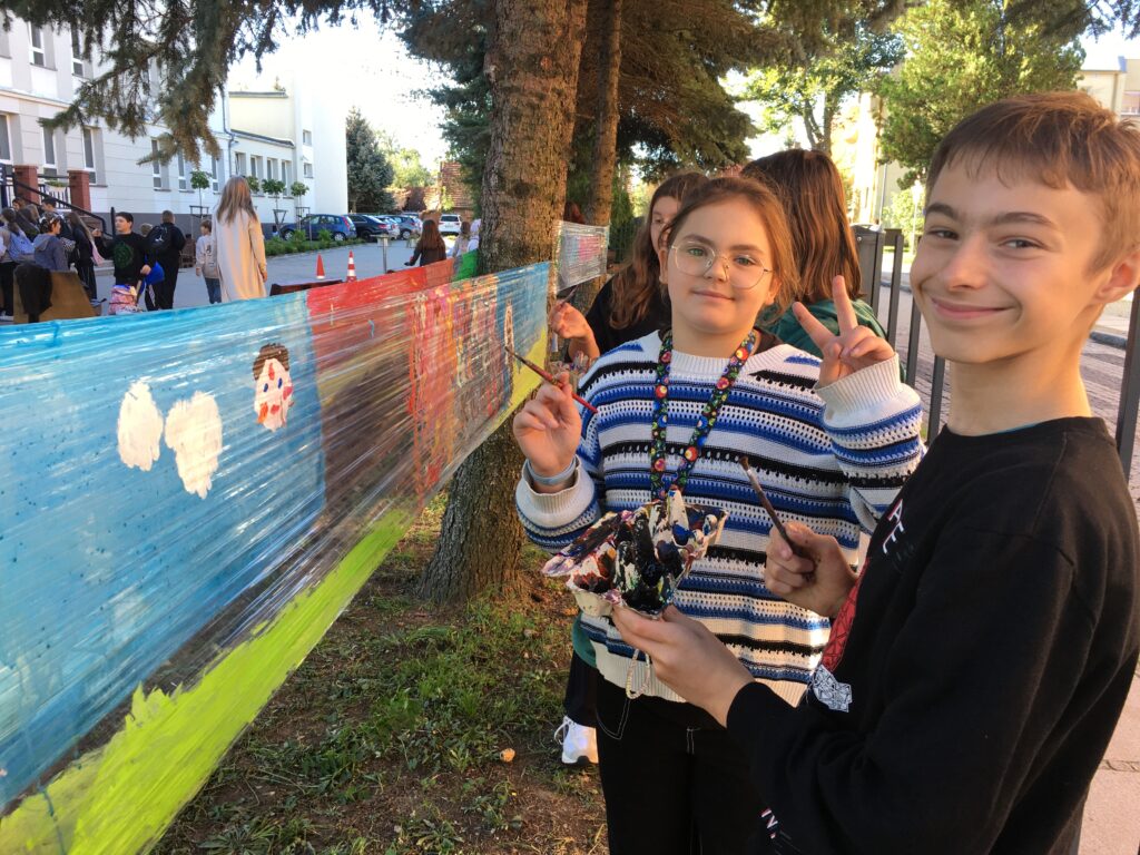 Podwórko szkolne, dziewczynka z chłopcem  na tle swojego dzieła stoją z rozłożonymi rączkami. Stoi na tle powierzchni folii, rozciągniętej między drzewami, po której maluje.