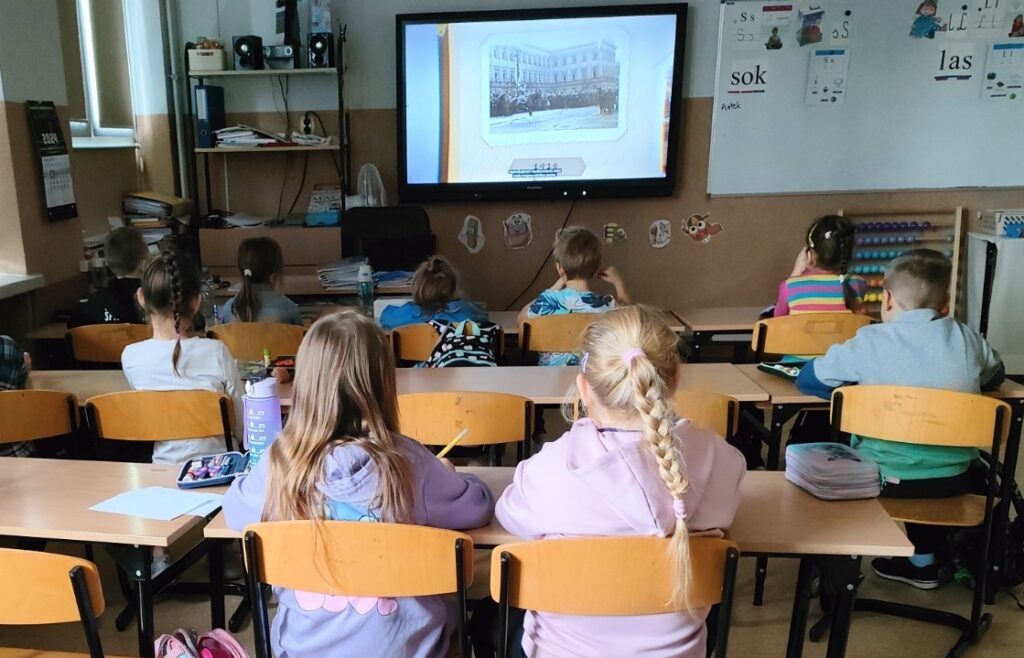  Dzieci siedzą w ławkach szkolnych i oglądają film wyświetlany na ekranie.